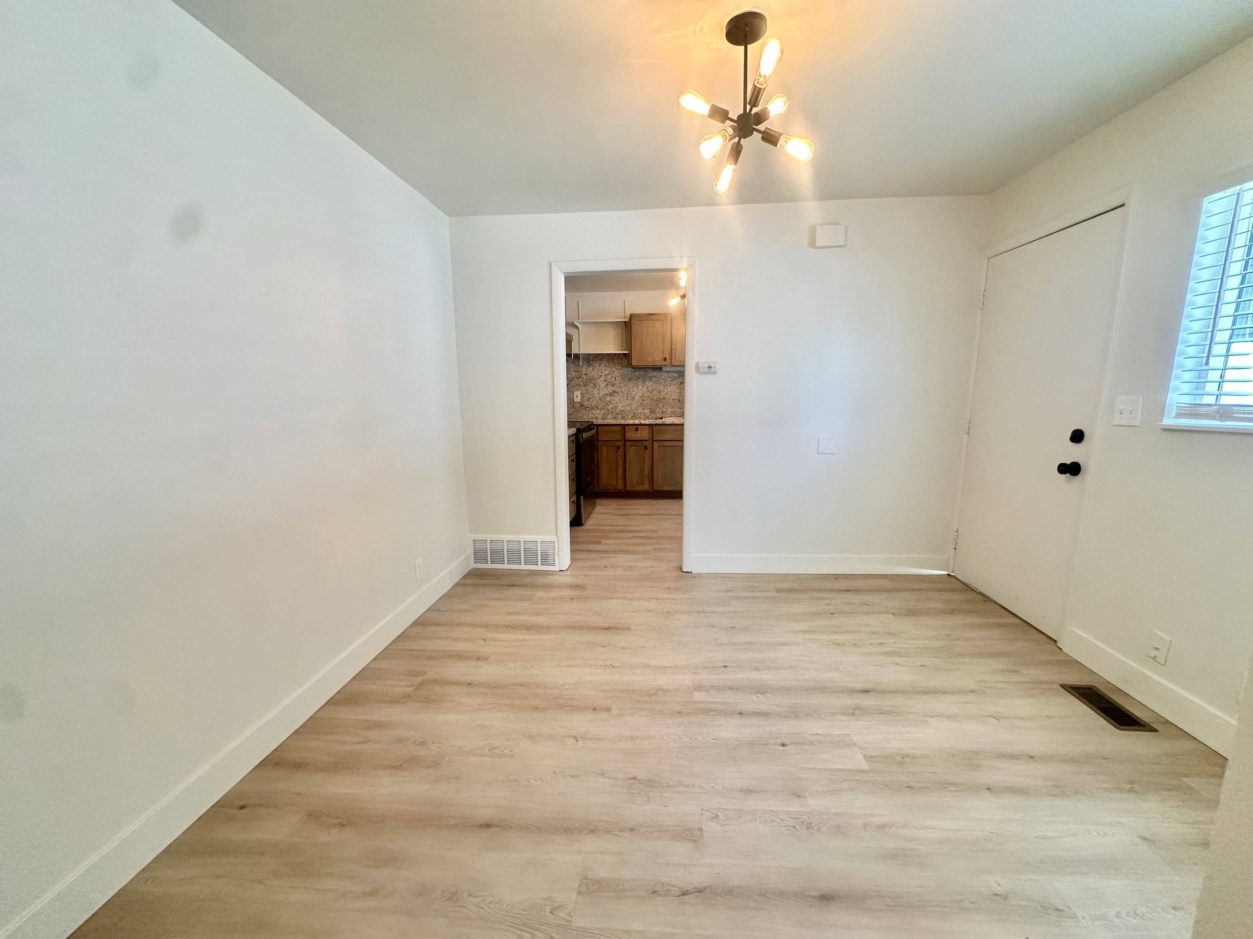 Open-concept area featuring light wood-finish flooring, a contemporary multi-bulb chandelier, white baseboards, and a white interior door with black hardware