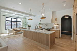 Kitchen featuring light wood finish cabinetry, arched walkways, light wood-type flooring, a center island with sink, and range hood