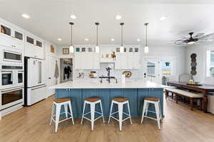 Two tone kitchen featuring pendant lighting, two tone color scheme, a large island with sink, glass fronted cabinets, and white appliances
