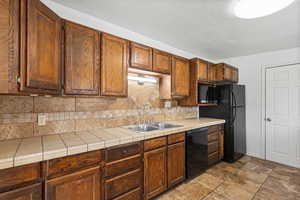 Kitchen featuring backsplash, tile countertops, black appliances, wood finish cabinets, and light stone finish floors