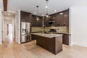 Kitchen with dark wood finish cabinetry, stainless steel appliances, an island with sink, light wood-type flooring, and pendant lighting