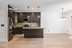 Kitchen featuring dark wood finish cabinets, stainless steel appliances, light wood-style flooring, a center island with sink, and backsplash