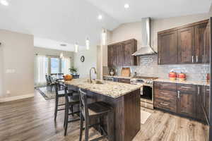 Kitchen with dark wood finish cabinetry, light stone counters, stainless steel range with gas stovetop, a kitchen breakfast bar, and vaulted ceiling