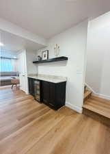 Indoor dry bar featuring dark cabinetry, light wood-style floors, open shelves, light countertops, and wine cooler