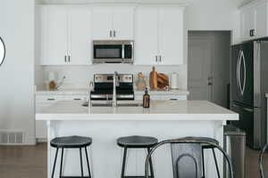 Kitchen featuring dark wood-type flooring, stainless steel appliances, white cabinetry, a kitchen island with sink, and a kitchen breakfast bar