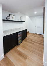 Indoor dry bar with dark cabinetry, open shelves, light wood-type flooring, and beverage cooler