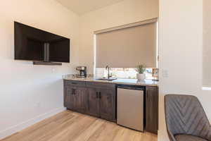 Indoor wet bar featuring dark wood finish cabinets, light wood-style floors, light stone counters, and dishwasher