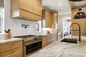 Kitchen featuring stainless steel appliances, light stone countertops, light wood finish cabinets, backsplash, and hanging light fixtures