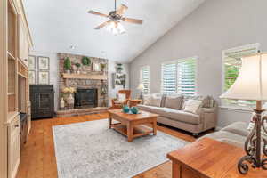 Living room featuring a brick fireplace, a ceiling fan, vaulted ceiling, and light wood-style flooring