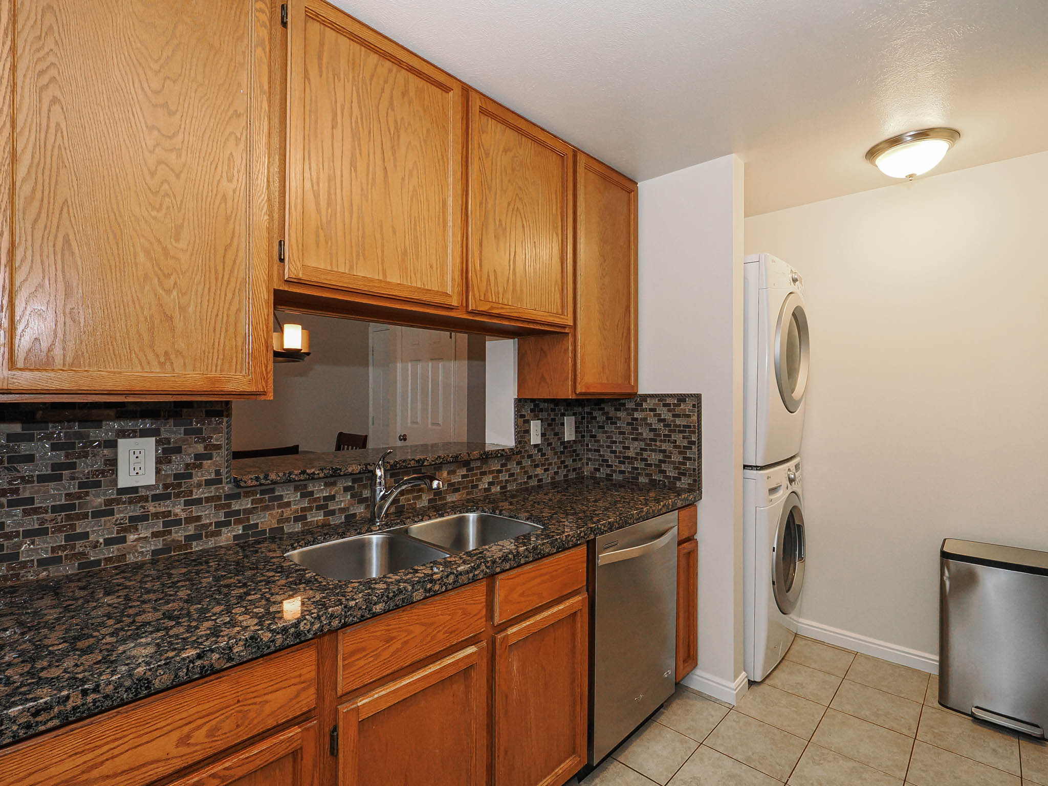 Kitchen with dishwasher, backsplash, dark stone counters, stacked washer and clothes dryer, and light tile patterned flooring