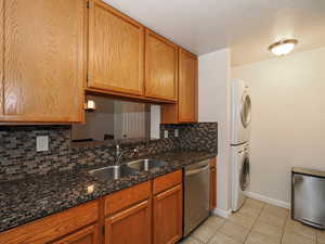 Kitchen with dishwasher, backsplash, dark stone counters, stacked washer and clothes dryer, and light tile patterned flooring