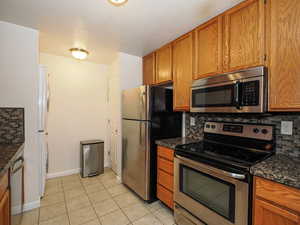 Kitchen featuring stainless steel appliances, tasteful backsplash, wood finish cabinets, light tile patterned floors, and dark stone counters