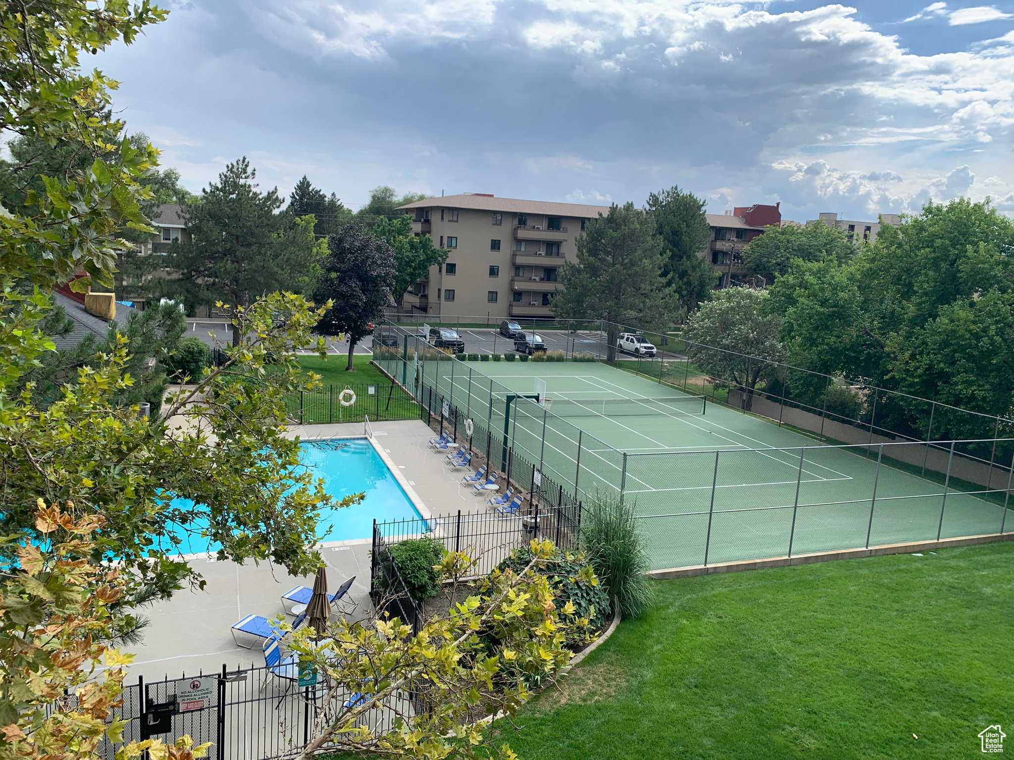 Expansive outdoor recreational area featuring a full-size tennis court with perimeter fencing, a rectangular swimming pool with concrete decking, and surrounding green spaces with mature trees