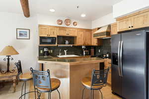 Kitchen with tile countertops, stainless steel fridge, decorative backsplash, black microwave, and light wood finish cabinets