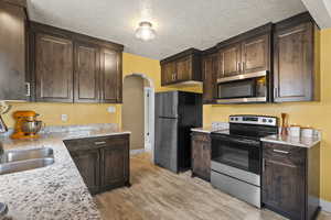 Kitchen featuring dark wood finish cabinets, stainless steel appliances, arched walkways, light stone countertops, and a textured ceiling