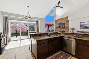 Kitchen featuring dark wood finish cabinetry, stainless steel dishwasher, a fireplace, dark stone countertops, and a chandelier
