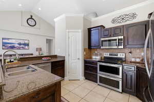 Kitchen featuring stainless steel appliances, dark stone countertops, dark wood finish cabinets, light tile patterned flooring, and ornamental molding