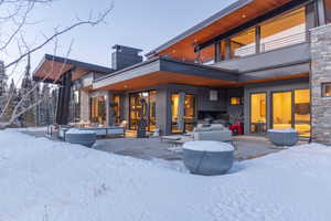 Snow covered rear of property with a patio area, stone siding, a chimney, and a balcony