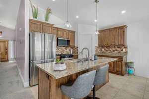 Kitchen with vaulted ceiling, light stone counters, stainless steel appliances, wood finish cabinets, and a kitchen island with sink