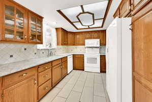 Kitchen with wood finish cabinetry, white appliances, glass fronted cabinets, light stone counters, and decorative backsplash