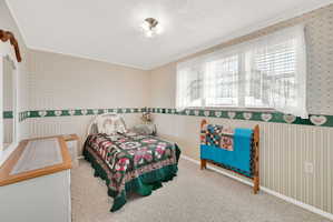 Bedroom with wallpapered walls, a wainscoted wall, ornamental molding, light colored carpet, and a textured ceiling