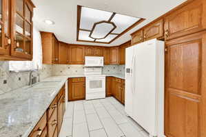 Kitchen featuring wood finish cabinets, light stone counters, white appliances, backsplash, and glass fronted cabinets