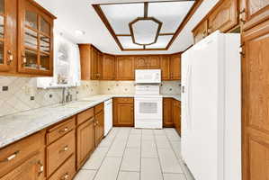 Kitchen featuring wood finish cabinetry, white appliances, light stone counters, glass fronted cabinets, and backsplash