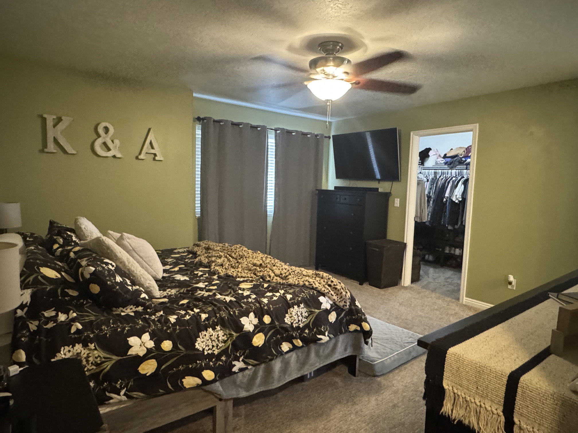 Carpeted bedroom featuring a ceiling fan with integrated lighting, window with drapes, and an open doorway revealing a walk-in closet with built-in shelving