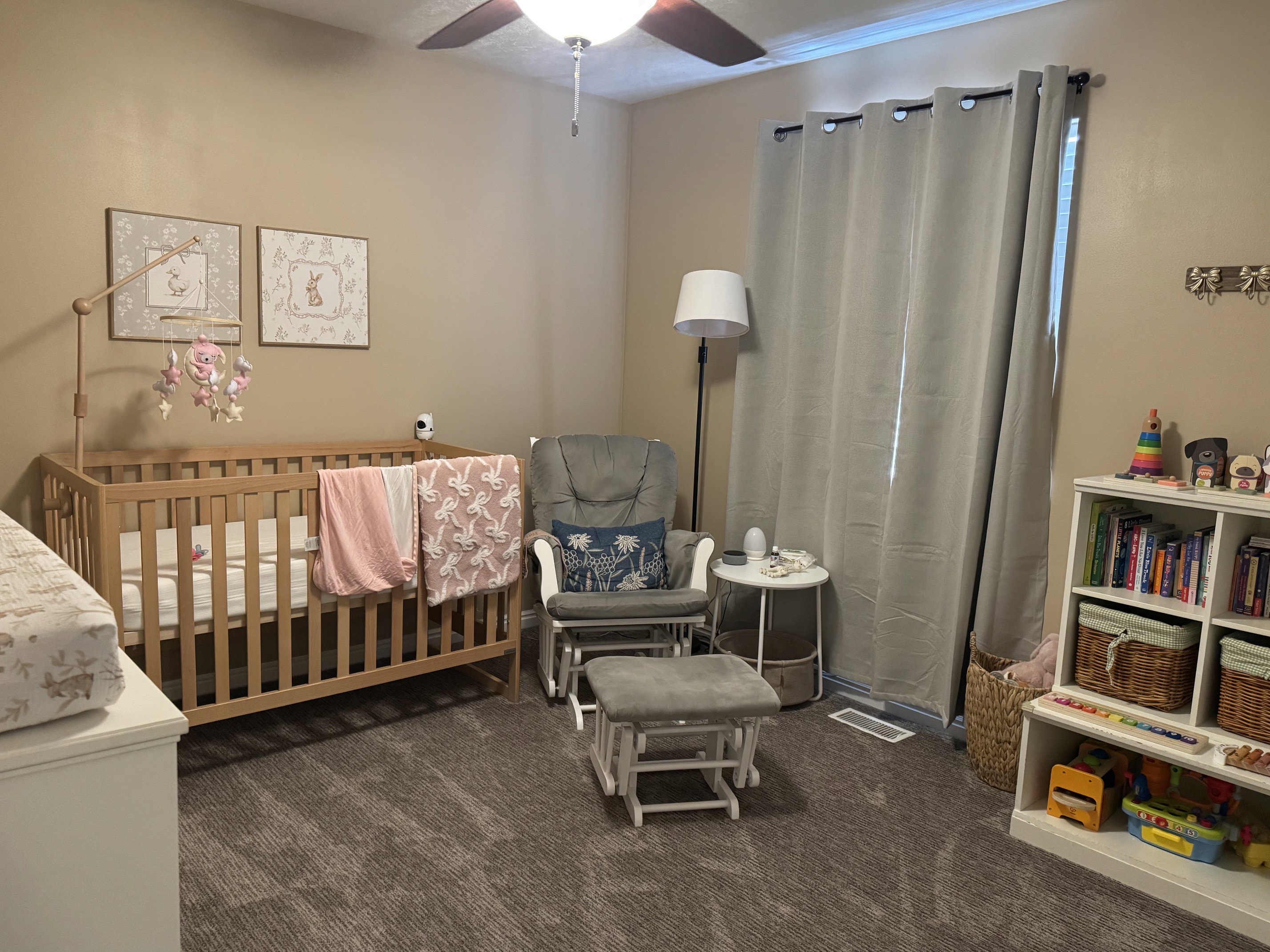 Carpeted room with a ceiling fan, featuring a light wood crib, a window with gray curtains, and a built-in white shelving unit