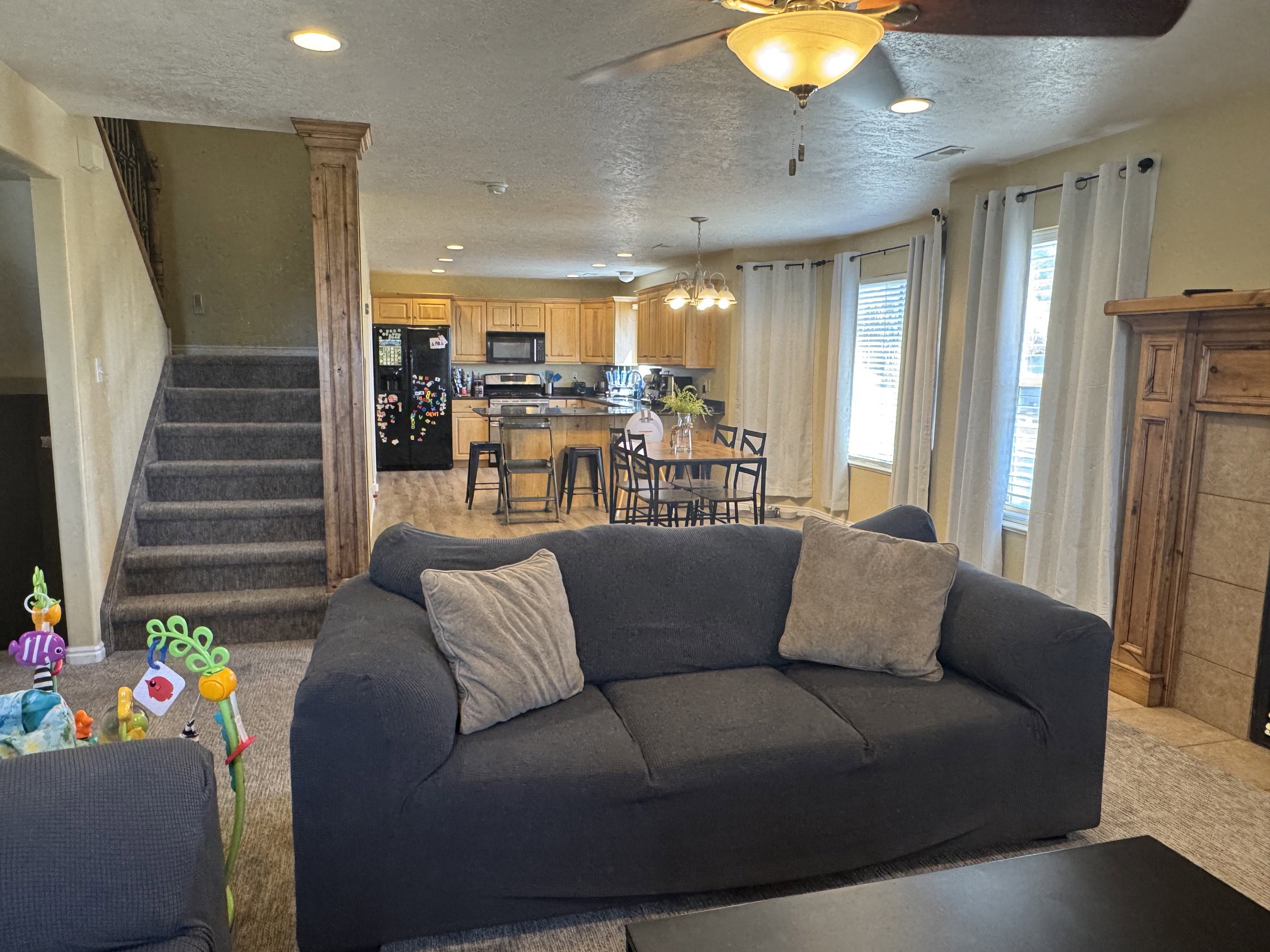 Open-concept living space featuring a carpeted staircase with wood-look newel post, a kitchen with wood cabinetry and dark appliances, a dining area with a circular table, and a living area with a built-in fireplace