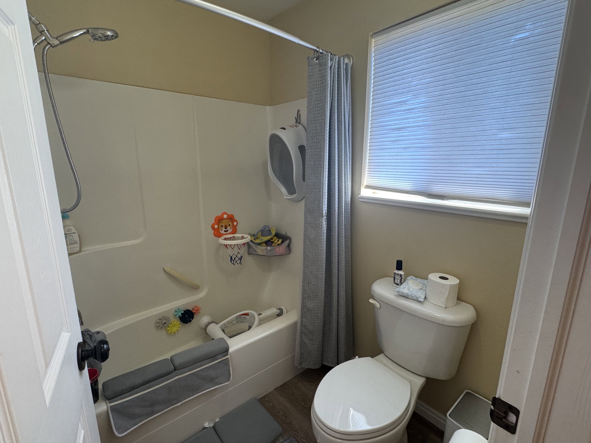 Full bathroom featuring a shower-over-tub combination with a handheld showerhead, a toilet, a window with privacy blinds, wood-finish flooring, and light-toned walls