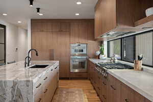 Kitchen with stainless steel appliances, light wood-style floors, light stone counters, recessed lighting, and a center island with sink