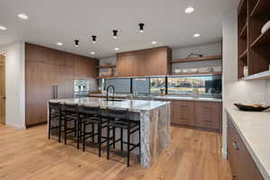 Kitchen with open shelves, light stone countertops, a large island with sink, wood finish cabinets, and recessed lighting