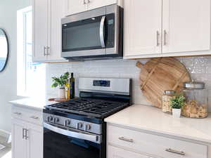 Example: Kitchen with stainless steel appliances, light stone counters, white cabinets, decorative backsplash, and plenty of natural light