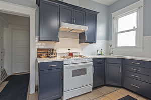 Kitchen featuring white range with gas stovetop, light tile patterned floors, light stone countertops, backsplash, and blue cabinetry