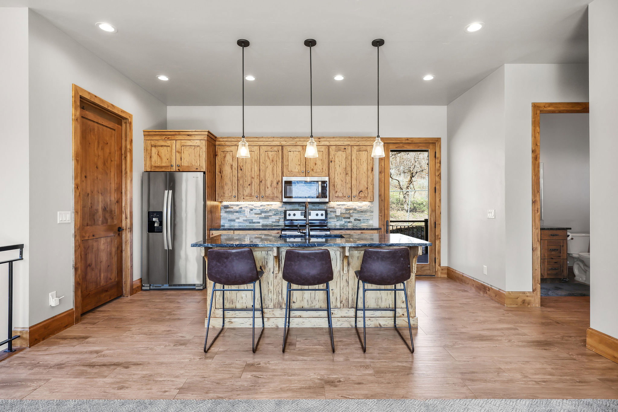 Kitchen with stainless steel appliances, a center island with sink, light wood-style flooring, decorative light fixtures, and dark stone countertops