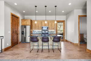 Kitchen with stainless steel appliances, a center island with sink, light wood-style flooring, decorative light fixtures, and dark stone countertops