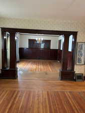 Unfurnished living room featuring ornate columns, light wood-type flooring, a chandelier, wallpapered walls, and wainscoting