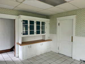 Kitchen featuring wainscoting, a drop ceiling, white cabinetry, wallpapered walls, and light tile patterned floors