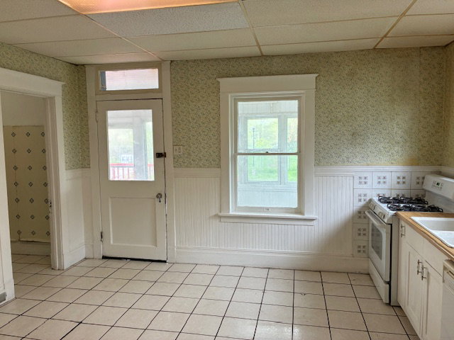Kitchen with wallpapered walls, white appliances, a paneled ceiling, wainscoting, and light countertops