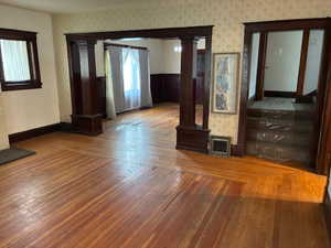 Unfurnished living room featuring ornate columns, wood-type flooring, wallpapered walls, and wainscoting