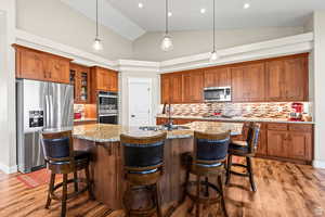 Kitchen with wood finish cabinets, stainless steel appliances, backsplash, light stone countertops, and vaulted ceiling
