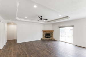 Unfurnished living room featuring a ceiling fan, a raised ceiling, dark wood-type flooring, a stone fireplace, and arched walkways
