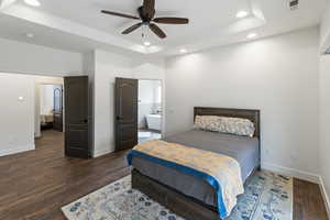 Bedroom featuring dark wood-style flooring, recessed lighting, ceiling fan, ensuite bathroom, and a tray ceiling