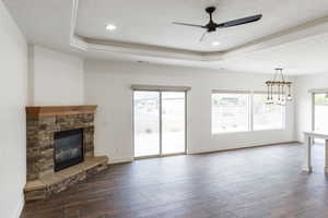 Unfurnished living room with plenty of natural light, a raised ceiling, dark wood-style flooring, a ceiling fan, and a stone fireplace