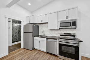 Kitchen with stainless steel appliances, light wood-style floors, dark stone counters, white cabinets, and vaulted ceiling