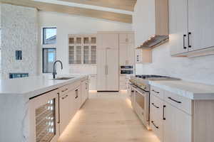 Kitchen with stainless steel appliances, light stone counters, wine cooler, light wood finish cabinetry, and lofted ceiling with beams