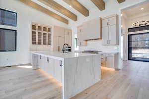 Kitchen featuring light wood finish cabinetry, light stone countertops, a center island with sink, light wood-style floors, and beam ceiling