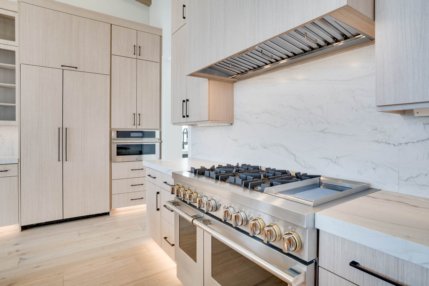 Kitchen with stainless steel appliances, light wood finish cabinetry, light wood-style flooring, light stone countertops, and decorative backsplash