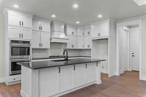 Kitchen featuring white cabinetry, stainless steel appliances, a center island with sink, tasteful backsplash, and dark wood-style floors